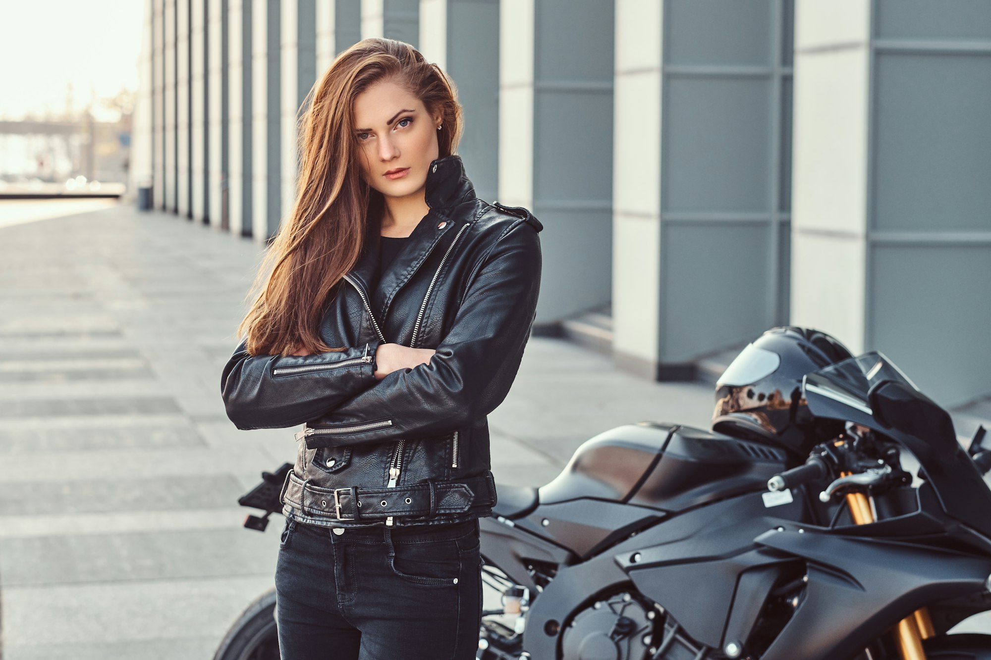 A confident biker girl with her arms crossed next to her superbike outside a building.